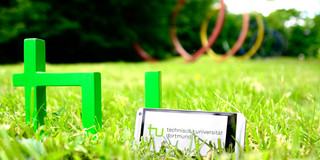 Ein Smartphone, das neben dem TU-Logo im Gras steht, im Hintergrund sind die Spektralringe zu sehen.